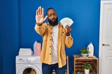 African american man holding money at laundry room with open hand doing stop sign with serious and confident expression, defense gesture