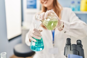Fototapeta premium Young blonde woman wearing scientist uniform pouring liquid on test tube at laboratory