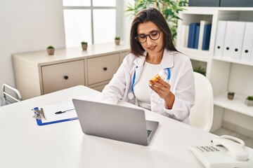 Young hispanic woman wearing doctor uniform holding pills working at clinic