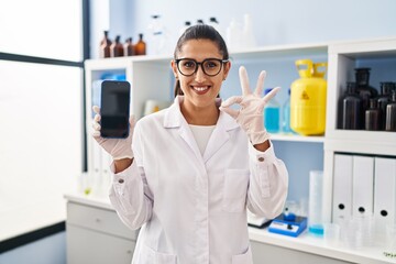 Young hispanic woman working at scientist laboratory with smartphone doing ok sign with fingers, smiling friendly gesturing excellent symbol