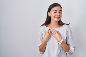 Young hispanic woman standing over white background smiling with hands on chest with closed eyes and grateful gesture on face. health concept.