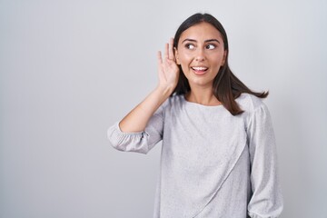 Young hispanic woman standing over white background smiling with hand over ear listening an hearing to rumor or gossip. deafness concept.