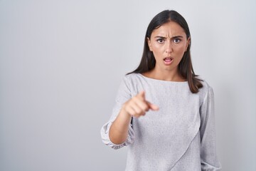 Young hispanic woman standing over white background pointing displeased and frustrated to the...