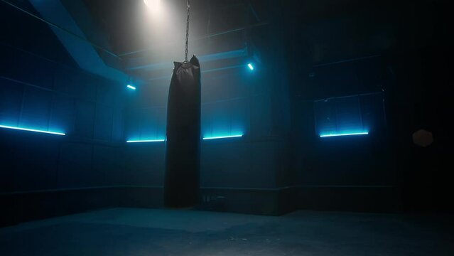 Long black punchbag hanging and swinging from chain in dark empty gym with blue neon lights. Leather punching bag for kickboxing, boxing, kicking and punching. Slow motion ready, 4K at 59.94fps.
