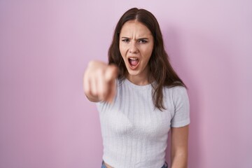 Young hispanic girl standing over pink background pointing displeased and frustrated to the camera,...