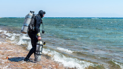 A man diver in scuba gear with one tank in hand and twin tanks enters the sea to start a technical dive