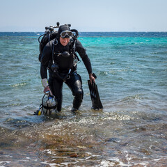 A smiling man diver in scuba gear with one cylinder in his right hand, fins in his left hand and twin cylinders comes out of the sea, after finishing a technical dive
