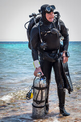 A satisfied smiling man diver in scuba gear with one cylinder in his right hand, fins in his left hand and twin cylinders stands on the seashore, after finishing a technical dive