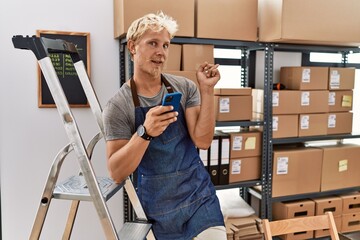 Young blond man using smartphone working at storehouse with a big smile on face, pointing with hand finger to the side looking at the camera.