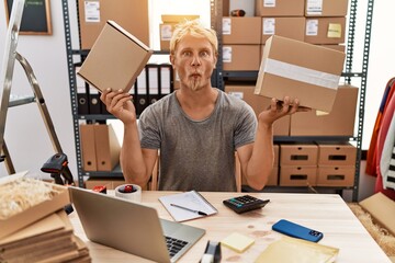 Young blond man holding packages working at online shop making fish face with mouth and squinting...