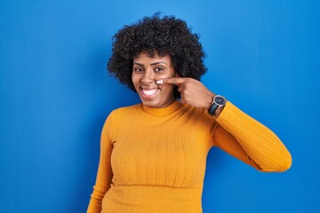 Fototapeta na wymiar Black woman with curly hair standing over blue background pointing with hand finger to face and nose, smiling cheerful. beauty concept