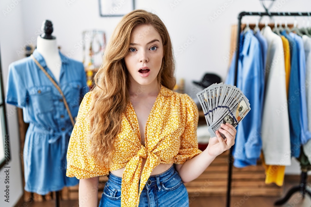 Poster young caucasian woman at retail boutique holding dollars banknotes scared and amazed with open mouth