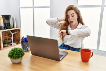 Young caucasian woman working at the office using computer laptop looking at the watch time worried, afraid of getting late