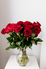 A beautiful bouquet of red roses in a glass vase stands on a white pedestal in a bright room. Red roses. Background with roses for Valentine's Day. Valentine's Day