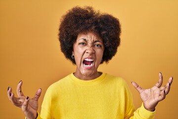 Young african american woman standing over yellow background crazy and mad shouting and yelling with aggressive expression and arms raised. frustration concept.