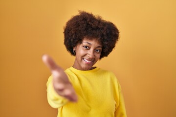 Obraz na płótnie Canvas Young african american woman standing over yellow background smiling friendly offering handshake as greeting and welcoming. successful business.