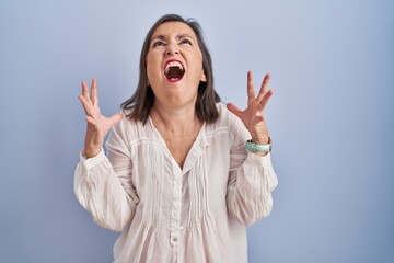 Middle age hispanic woman standing over blue background crazy and mad shouting and yelling with aggressive expression and arms raised. frustration concept.