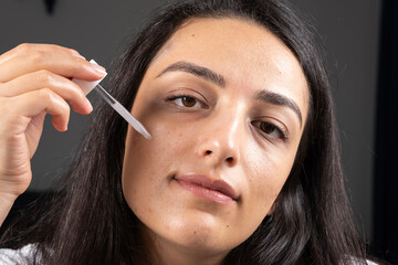 Obraz premium Facial skin care, brunette woman looking camera while facial skin care. Using, applying serum her face. Moisturizing or acne treatment. Modern, attractive girl beauty routine concept idea, copy space.