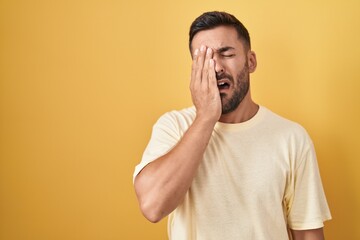 Handsome hispanic man standing over yellow background yawning tired covering half face, eye and mouth with hand. face hurts in pain.