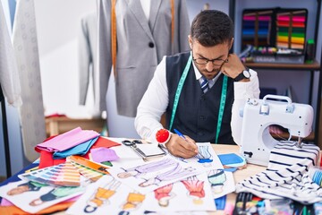 Young hispanic man tailor drawing clothing design on notebook at tailor shop