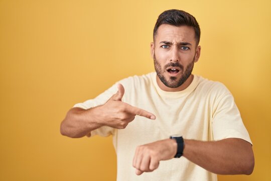 Handsome hispanic man standing over yellow background in hurry pointing to watch time, impatience, upset and angry for deadline delay