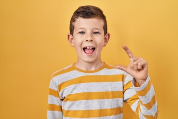 Young caucasian kid standing over yellow background smiling and confident gesturing with hand doing small size sign with fingers looking and the camera. measure concept.