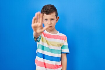 Young caucasian kid standing over blue background doing stop sing with palm of the hand. warning expression with negative and serious gesture on the face.