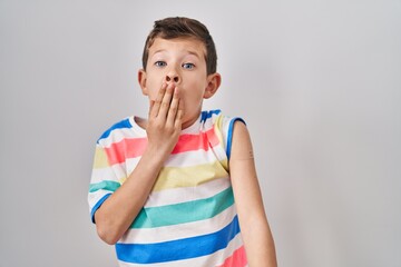 Young caucasian kid getting vaccine showing arm with band aid covering mouth with hand, shocked and...