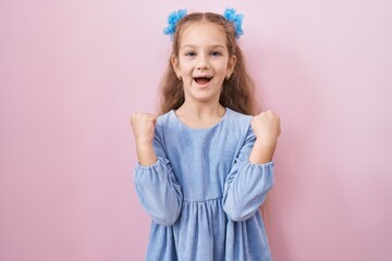 Young little girl standing over pink background screaming proud, celebrating victory and success very excited with raised arms