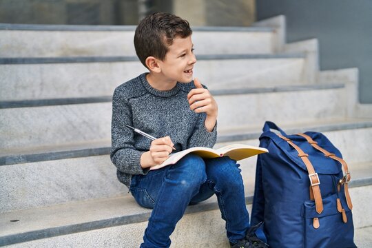 Blond child student writing on book sitting on stairs at school