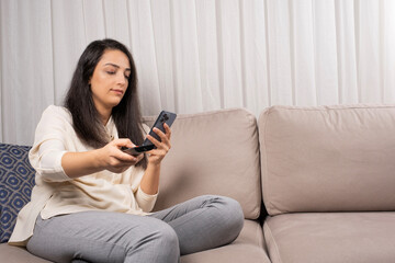 Smartphone and social media addiction, caucasian brunette woman holding tv remote, looking smartphone and social media addiction. Sitting couch at home want to watch television. Zapping concept idea.