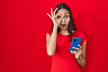 Naklejka premium Young brazilian woman using smartphone over red background doing ok gesture shocked with surprised face, eye looking through fingers. unbelieving expression.