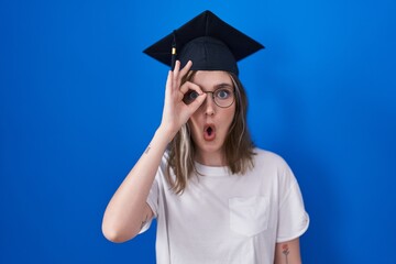 Blonde caucasian woman wearing graduation cap doing ok gesture shocked with surprised face, eye looking through fingers. unbelieving expression.