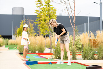 Mother playing with her cute daughter mini golf at outdoor playground.