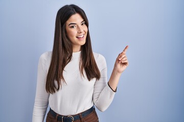 Young brunette woman standing over blue background with a big smile on face, pointing with hand finger to the side looking at the camera.