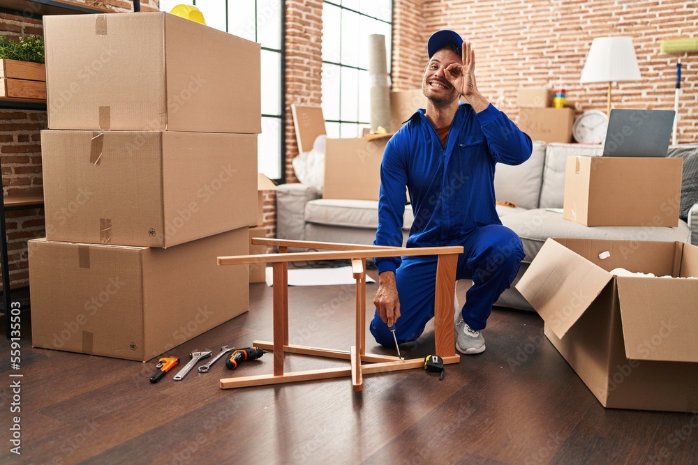 Poster Young hispanic man working on moving service smiling happy doing ok sign with hand on eye looking through fingers