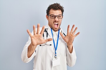 Fototapeta na wymiar Young hispanic man wearing doctor uniform and stethoscope afraid and terrified with fear expression stop gesture with hands, shouting in shock. panic concept.