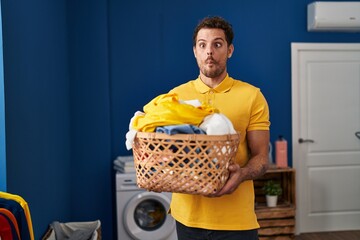 Young hispanic man holding laundry basket making fish face with mouth and squinting eyes, crazy and...