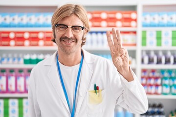 Caucasian man with mustache working at pharmacy drugstore showing and pointing up with fingers number three while smiling confident and happy.