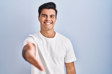 Hispanic man standing over blue background smiling friendly offering handshake as greeting and welcoming. successful business.
