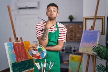 Brazilian young man holding painter palette at artist studio puffing cheeks with funny face. mouth...