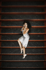 Obraz premium Lush hair volume curly hair; A dark-haired girl is dressed in a light elegant dress; a sexy girl lies on the steps of a building; red lipstick on the lips;