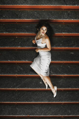 Obraz premium Lush hair volume curly hair; A dark-haired girl is dressed in a light elegant dress; a sexy girl lies on the steps of a building; red lipstick on the lips;