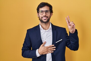 Handsome latin man standing over yellow background smiling swearing with hand on chest and fingers...