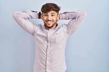 Arab man with beard standing over blue background relaxing and stretching, arms and hands behind head and neck smiling happy