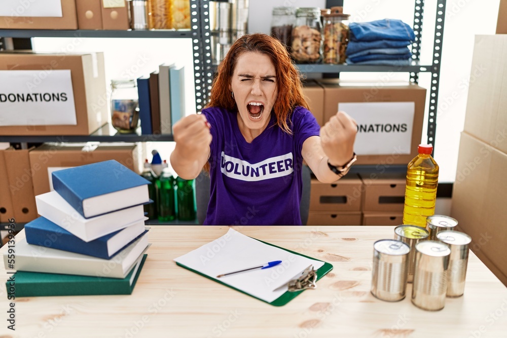 Poster Young redhead woman wearing volunteer t shirt at donations stand angry and mad raising fists frustrated and furious while shouting with anger. rage and aggressive concept.