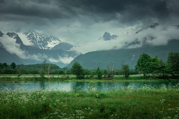Landscape of Romsdalen with snowy mountains of Troll Wall in rain clouds and flowers with river in foreground, in Norway