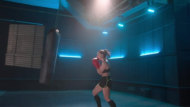 Kickboxing training in dark gym. Woman fighter trains punches and kicks with trainer in sparring. Female athlete in boxing gloves is practicing hitting punching bag. Slow motion ready, 4K at 59.94fps.