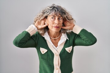 Middle age woman standing over white background trying to hear both hands on ear gesture, curious...
