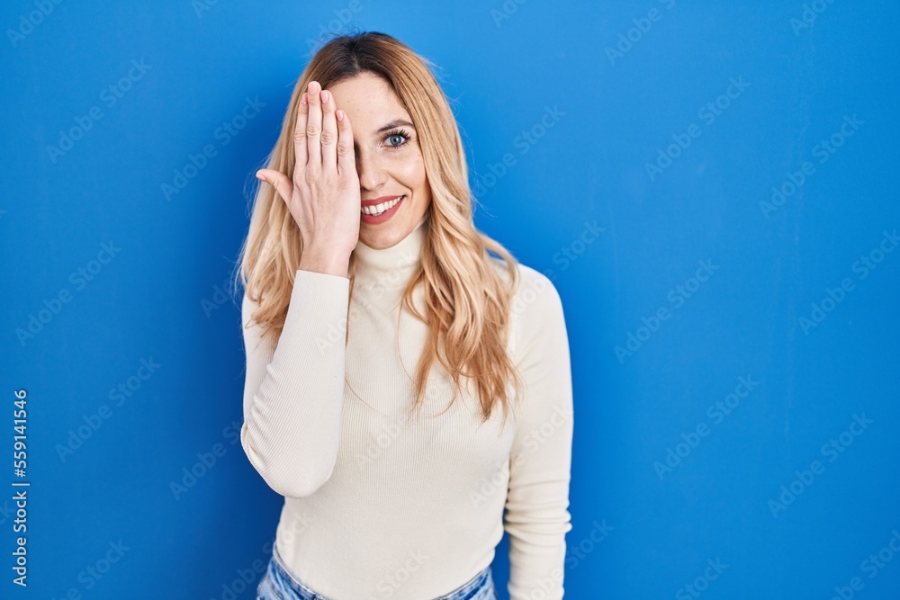 Sticker Young caucasian woman standing over blue background covering one eye with hand, confident smile on face and surprise emotion.